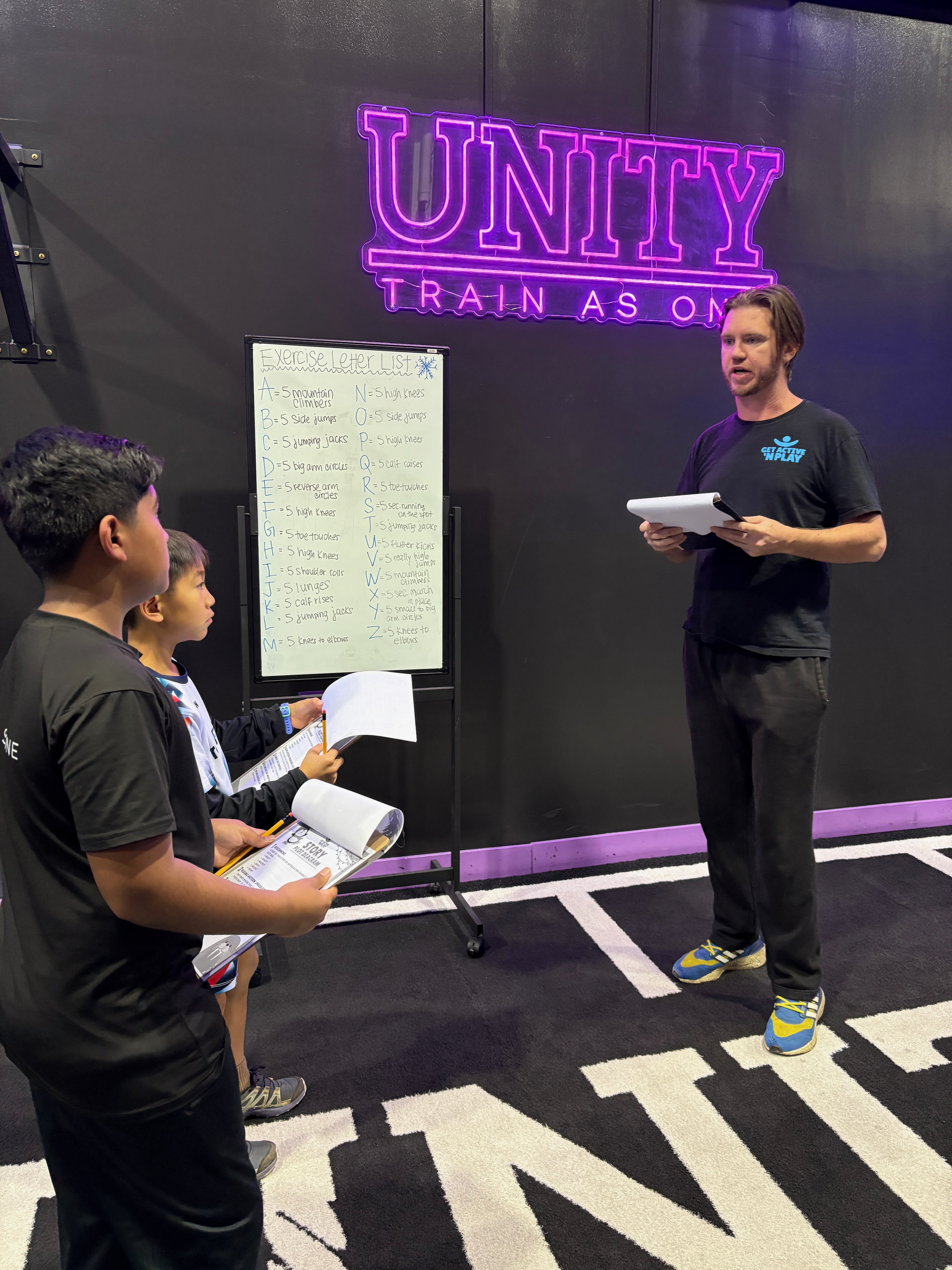 A GANP coach explains the Story Challenge to two kids holding clipboards at UNITY